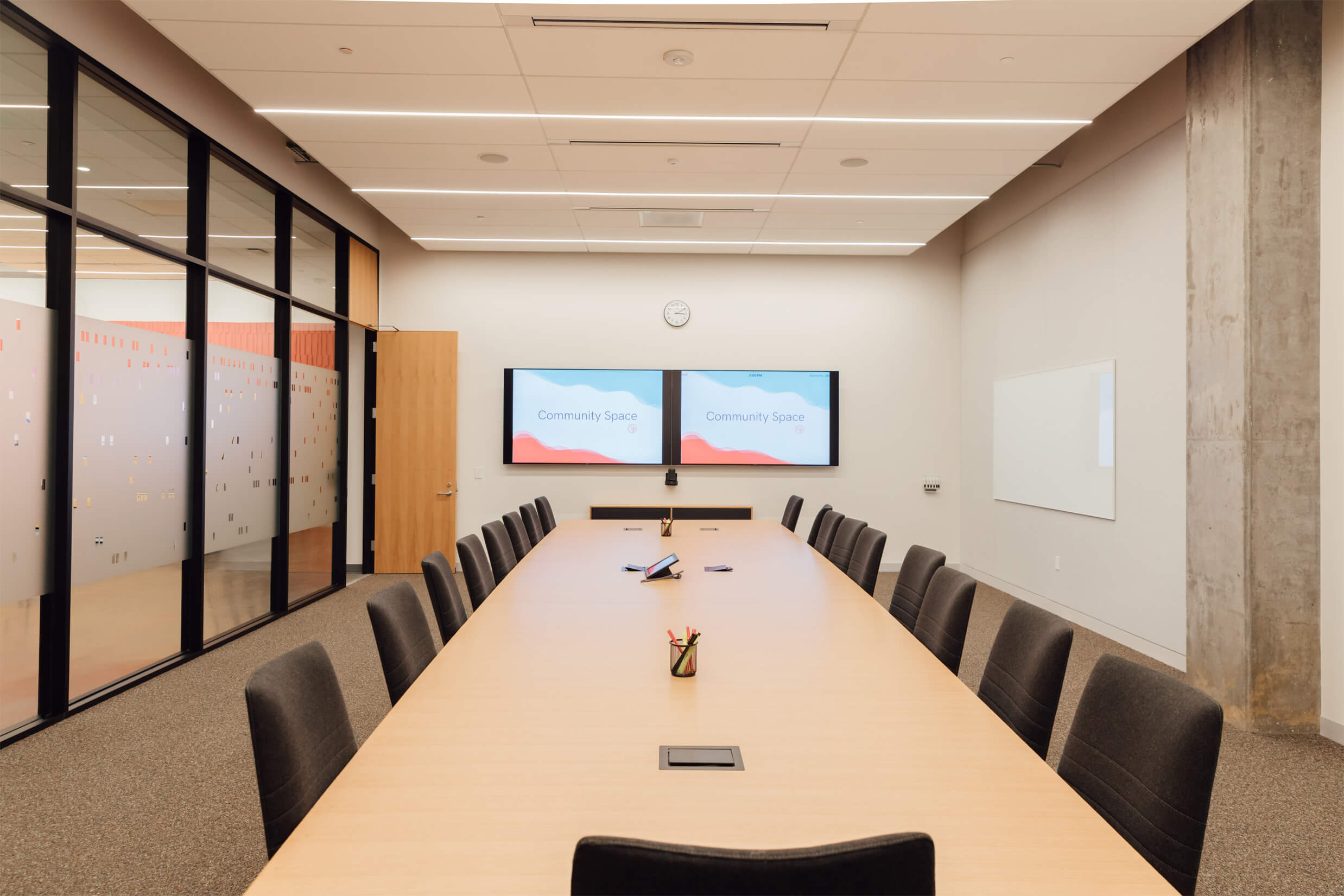 Large conference room with a table and 17 chairs, facing two digital screens.