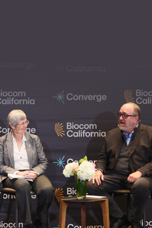 Sandy Schmid and Marc Malandro speak on stage at a Biocom California Converge event, seated with a flower arrangement between them.
