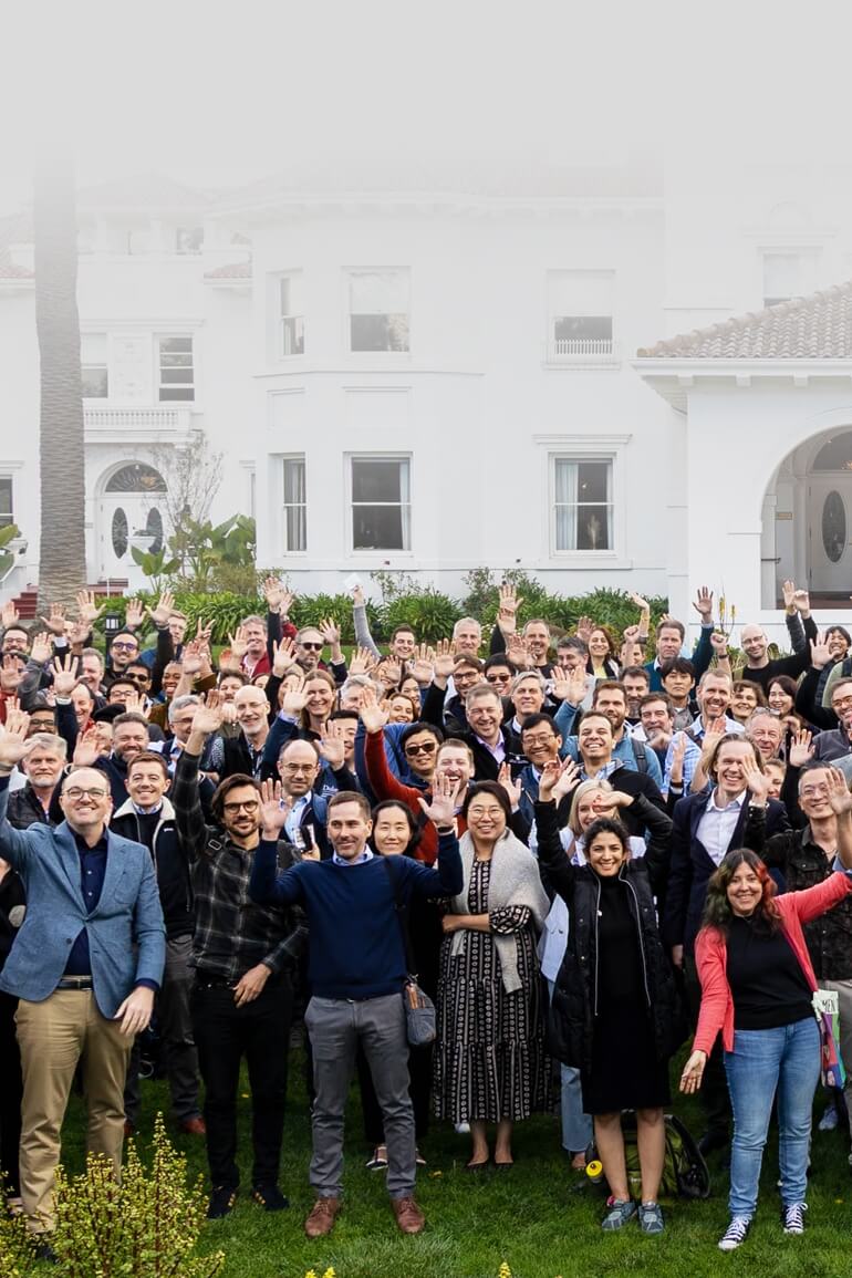 A large group of people stands outdoors under tall trees, smiling and waving at the camera.